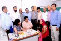 Indian mother and two children talks to seated man and woman in a clinical setting, with group of men standing around them.
